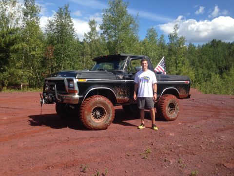 1979 Ford Bronco - A man stands next to a lifted, muddy black Ford pickup truck with an American flag in the bed, parked on a dirt road with trees in the back…