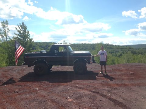 1979 Ford Bronco - A man stands next to a dark colored Ford Bronco with an American flag mounted on the rear. The vehicle is parked on a dirt road with a wood…