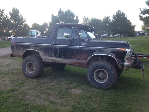 1979 Ford Bronco - A black Ford Bronco is parked on a grassy area, covered in mud splatters. The vehicle is viewed from the side, with large off-road tires an…