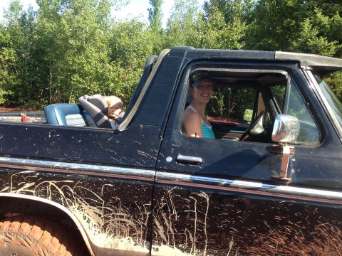 1979 Ford Bronco - A woman smiles from the driver's seat of a muddy black Ford Bronco, with a child resting in a car seat in the passenger area. The vehicle's…