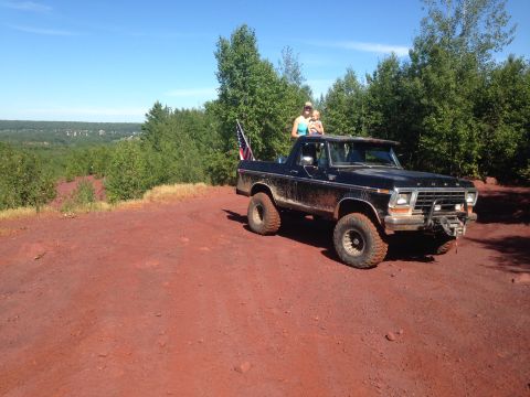 1979 Ford Bronco - A black 1979 Ford Bronco with an American flag is parked on a dirt road overlooking a wooded valley. Two people are standing in the open to…