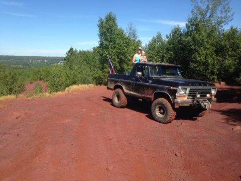 1979 Ford Bronco - A black 1979 Ford Bronco is parked on a dirt road with a woman and child standing on top. An American flag is attached to the rear of the v…