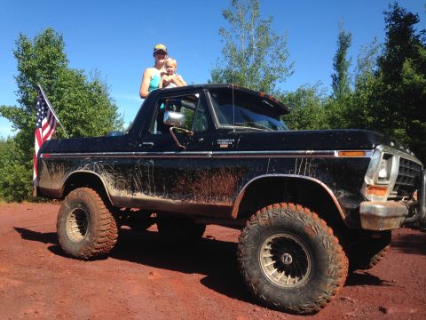 1979 Ford Bronco - A black Ford Bronco is parked on a dirt road with an American flag in the bed. A woman and child are standing in the passenger seat area.