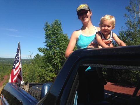 1979 Ford Bronco - A woman and a young child are standing in the bed of a dark-colored pickup truck, with an American flag mounted on the side. The truck is p…