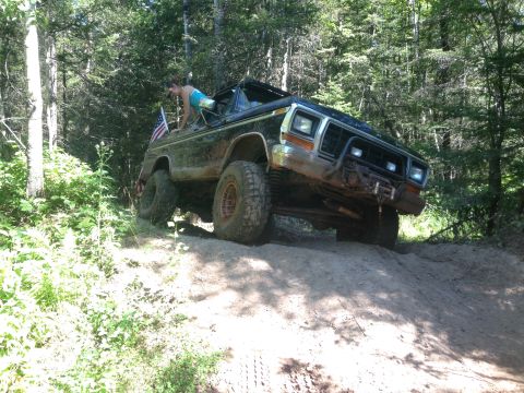 1979 Ford Bronco - A person in a blue top is climbing out of a black Ford Bronco that is driving up a dirt hill in a wooded area. An American flag is attached…