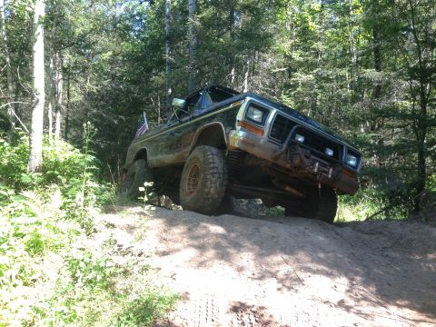 1979 Ford Bronco - A dark-colored Ford Bronco is shown from a low angle, driving up a dirt hill in a wooded area. The vehicle has large off-road tires and a b…