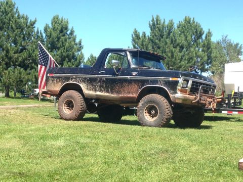 1979 Ford Bronco - A black, lifted 1979 Ford Bronco covered in mud sits on a grassy field under a clear blue sky. An American flag is mounted on the rear of t…