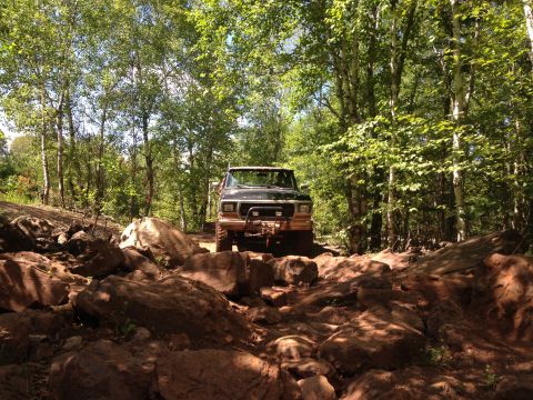 1979 Ford Bronco - A dark colored Ford Bronco is navigating a rocky, off-road trail surrounded by trees. The front of the vehicle is visible, equipped with a…