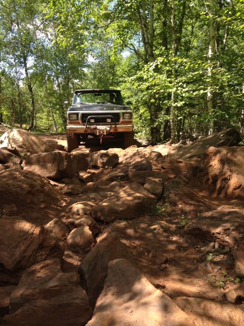1979 Ford Bronco - A black Ford Bronco is navigating a rocky, off-road trail surrounded by trees. The vehicle has a winch mounted on the front bumper.