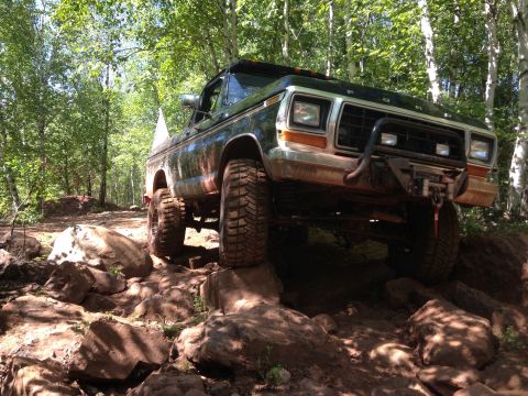 1979 Ford Bronco - A dark colored Ford Bronco is navigating a rocky, wooded trail. The vehicle is angled to the right, showcasing its off-road tires and winch.