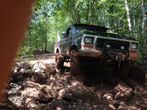 1979 Ford Bronco - A dark colored Ford Bronco is navigating a rocky, wooded trail. The front of the vehicle shows a bull bar and a winch.