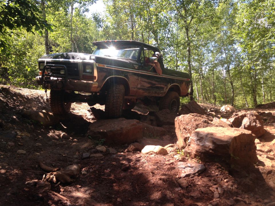 Lifted dark Ford Bronco off-roading on a rocky, wooded trail.