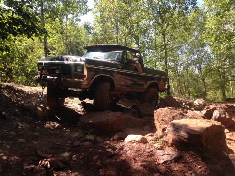 1979 Ford Bronco - A dark-colored, lifted 1970s Ford Bronco navigates a rocky, wooded trail. The vehicle is angled towards the viewer, showing its front and s…