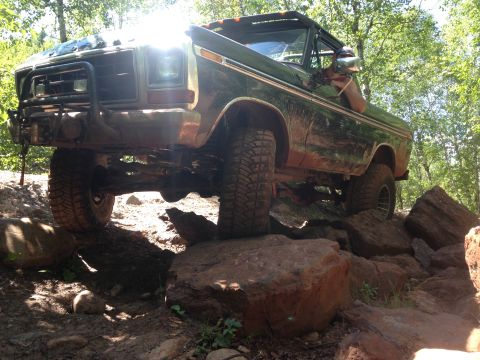 1979 Ford Bronco - A dark green, lifted Ford Bronco is navigating a rocky off-road trail, with its front tire on a large boulder. The sun glints off the winds…