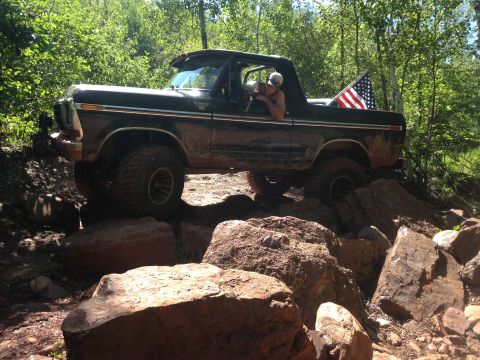 1979 Ford Bronco - A dark-colored, lifted Ford Bronco is navigating a rocky, wooded trail. An American flag is mounted on the back of the vehicle.