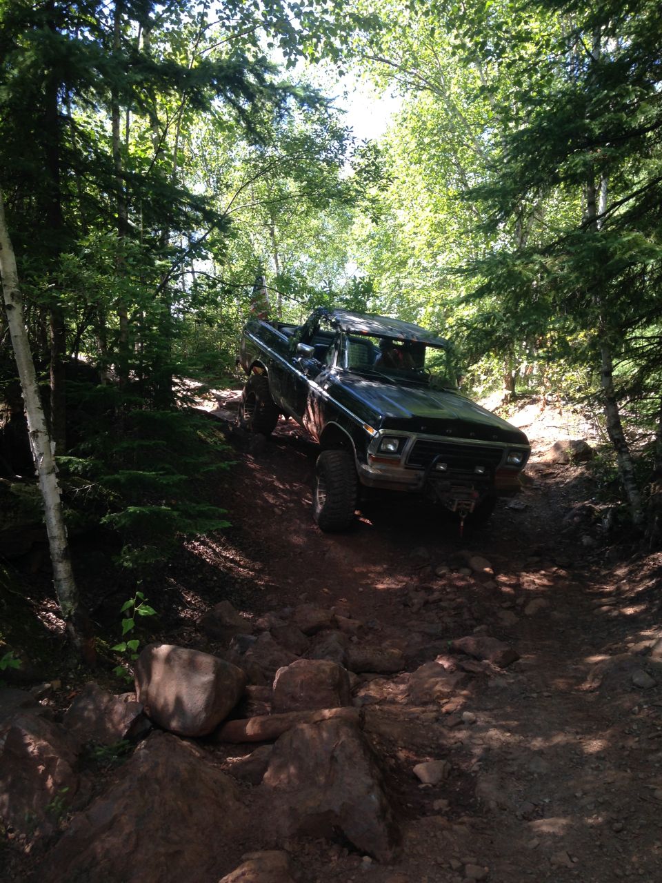 1979 Ford Bronco with top removed driving on a rocky trail in a forest.