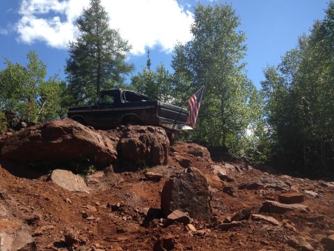 1979 Ford Bronco - A black 1979 Ford Bronco is parked on a rocky, red dirt hill with trees and a blue sky in the background. An American flag is attached to t…