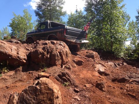 1979 Ford Bronco - A dark colored Ford Bronco is navigating a steep, rocky, and muddy off-road trail. An American flag is attached to the rear of the vehicle.