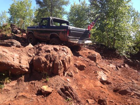 1979 Ford Bronco - A dark-colored, two-door SUV with an American flag attached to the rear is navigating a steep, rocky, and muddy off-road trail.