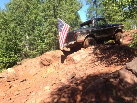1979 Ford Bronco - A black 1979 Ford Bronco is shown off-roading on a steep, rocky, and muddy incline. An American flag is attached to the rear of the vehicle.