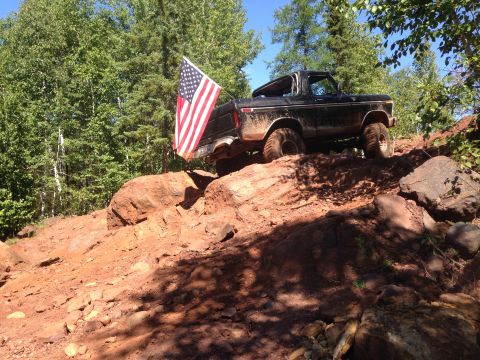 1979 Ford Bronco - A black 1979 Ford Bronco is shown driving up a rocky, dirt incline with an American flag attached to the rear. The vehicle is partially obs…