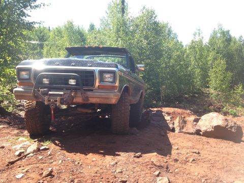 1979 Ford Bronco - A black 1979 Ford Bronco is shown from the front three-quarter view, navigating a rocky, dirt trail surrounded by trees. The vehicle featur…
