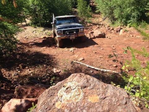1979 Ford Bronco - A black 1979 Ford Bronco is navigating a rocky, off-road trail surrounded by trees. The truck has an American flag attached to the antenna…