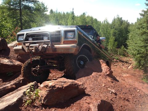 1979 Ford Bronco - A dark-colored Ford Bronco is navigating a rocky, off-road trail with its front tires high on a mound of dirt and rocks. The driver is visi…