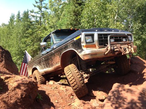 1979 Ford Bronco - A black, lifted 1979 Ford Bronco is navigating a rocky, off-road trail. An American flag is mounted on the driver's side rear.