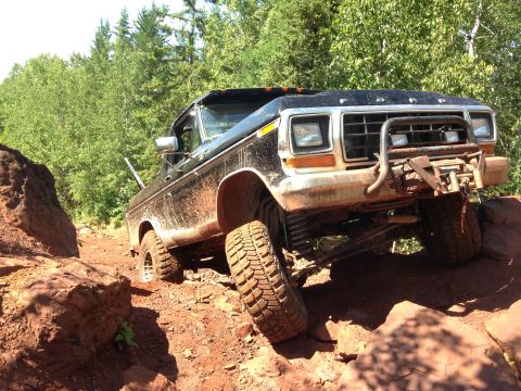 1979 Ford Bronco - A black 1979 Ford Bronco is shown from a low 3/4 front angle, navigating a rocky, off-road trail with red dirt and trees in the background.…