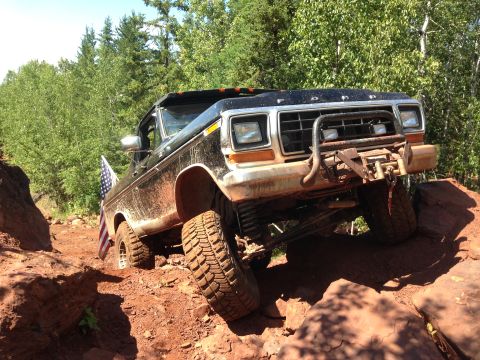 1979 Ford Bronco - A black 1979 Ford Bronco is shown off-roading on a rocky, dirt trail with an American flag attached to the driver's side. The vehicle is ti…