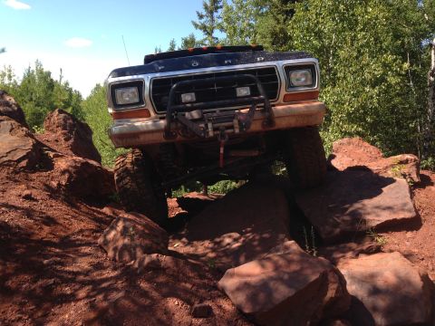 1979 Ford Bronco - A black Ford truck is navigating a rocky, off-road trail with large boulders and red dirt. The front of the truck, including its grille, he…
