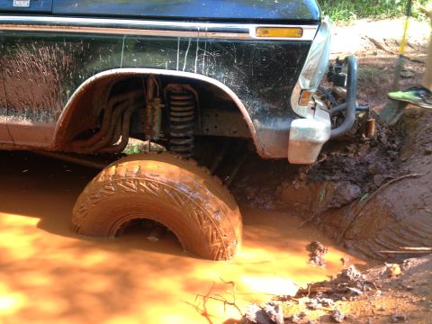 1979 Ford Bronco - A black Ford Bronco is stuck in deep mud, with its front passenger-side tire submerged. The vehicle's suspension and exhaust system are vis…