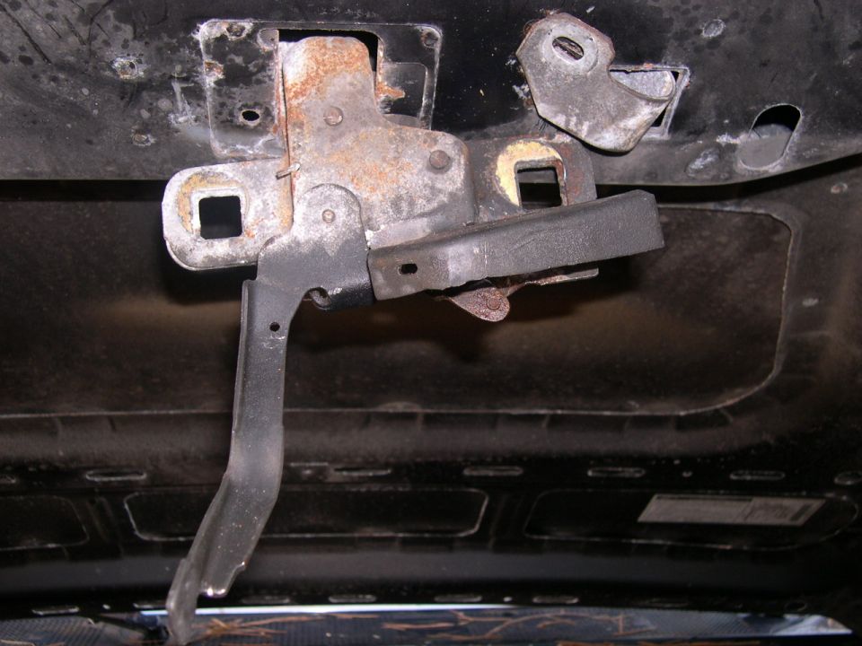 Underside view of a tailgate or hatch latch mechanism with rusty striker and metal linkage