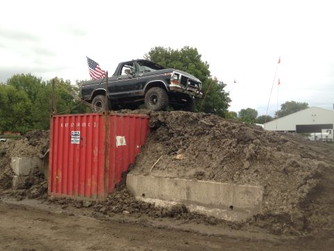 1979 Ford Bronco - A black 1979 Ford Bronco is parked on a dirt mound next to a red shipping container, with an American flag flying from the truck bed. The v…