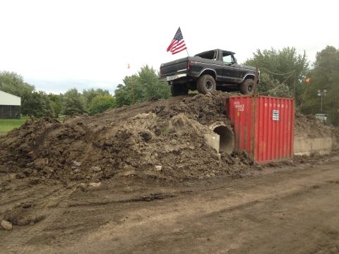 1979 Ford Bronco - A black 1979 Ford Bronco with an American flag mounted on the tailgate is positioned on a large dirt mound, partially obscuring a red shipp…