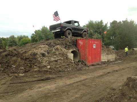 1979 Ford Bronco - A black Ford Bronco with an American flag on the tailgate is parked on a dirt mound, partially obscuring a large red shipping container and…