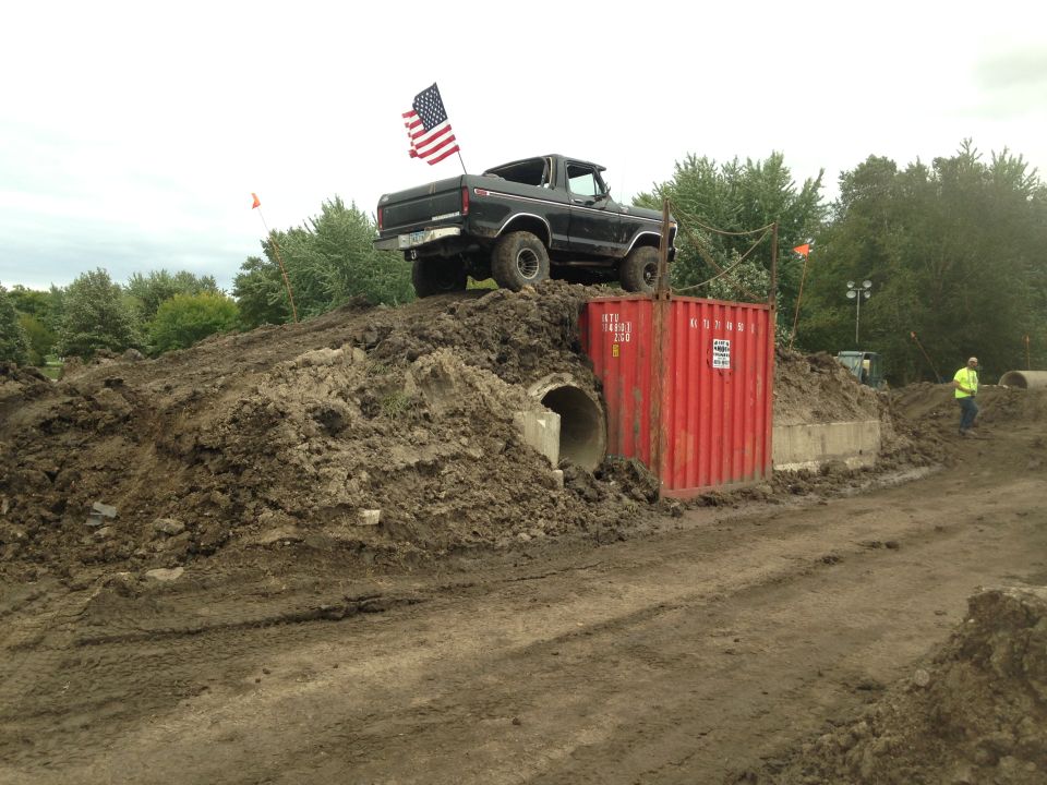 Black Ford Bronco with American flag on dirt mound next to red shipping container.