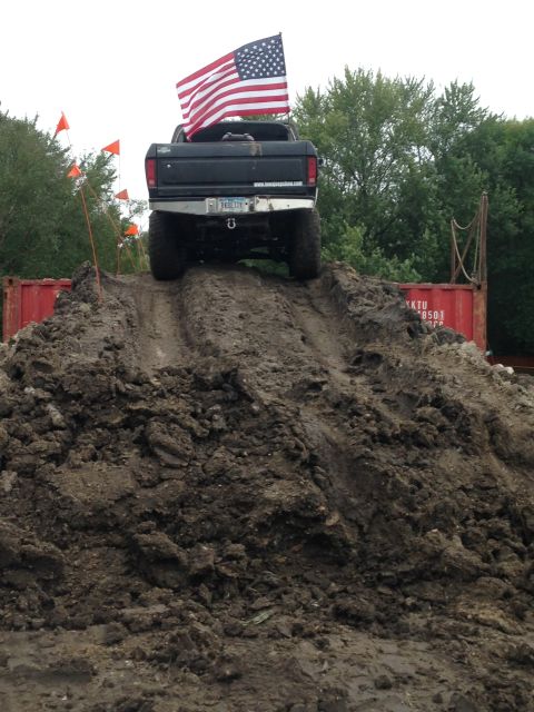 1979 Ford Bronco - The rear of a black Ford pickup truck is shown ascending a steep dirt mound, flying an American flag from its bed. The truck has large tire…