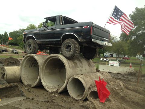1979 Ford Bronco - A black 1979 Ford Bronco is positioned on a dirt obstacle course, with its front tires resting on large concrete pipes. An American flag is…
