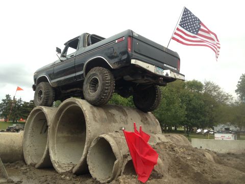 1979 Ford Bronco - A black 1979 Ford Bronco is positioned on top of large concrete pipes, with an American flag flying from its tailgate. The vehicle is viewe…