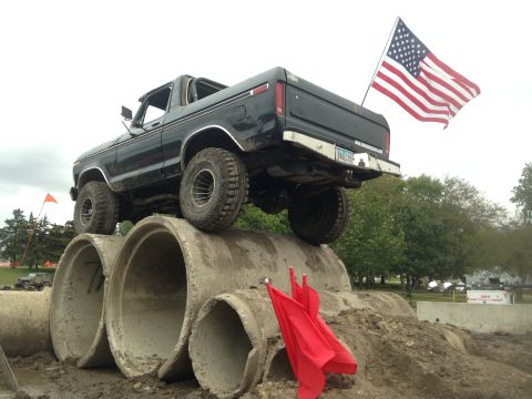 1979 Ford Bronco - A black Ford Bronco is positioned on top of large concrete pipes, with an American flag mounted on the rear. Two red flags are visible in t…