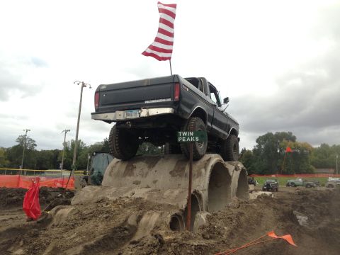 1979 Ford Bronco - A black 1970s Ford pickup truck is positioned on top of large concrete pipes, with an American flag waving from its bed. The truck has larg…