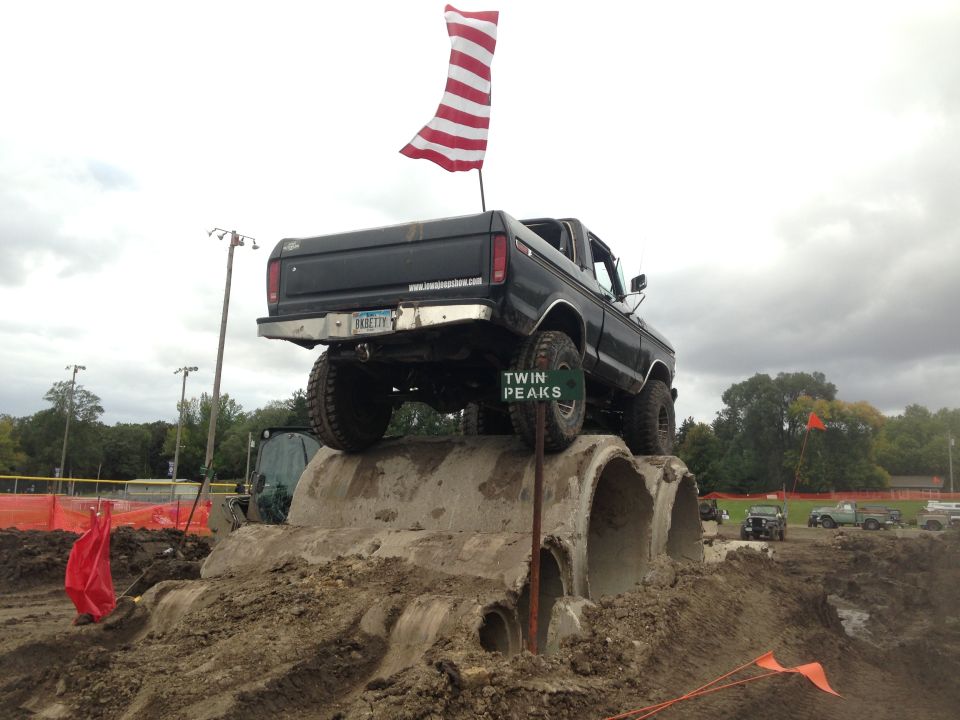 Black 1970s Ford pickup truck on concrete pipes with American flag.