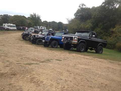 1979 Ford Bronco - A line of off-road vehicles, including a black Ford F-Series truck, are parked on a dirt lot with trees in the background.