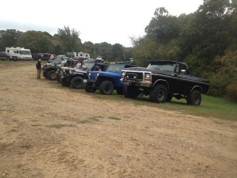 1979 Ford Bronco - A line of off-road vehicles, including a black 1979 Ford Bronco, parked on a dirt lot.