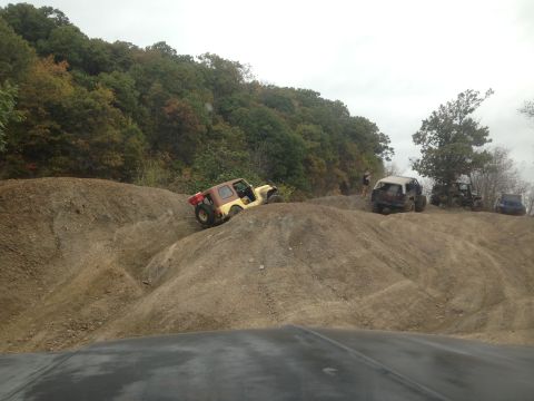 1979 Ford Bronco - Multiple off-road vehicles are seen traversing a challenging dirt terrain with wooded hills in the background.
