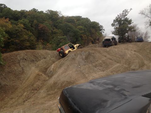1979 Ford Bronco - Multiple off-road vehicles traverse a challenging dirt terrain with wooded hills in the background.