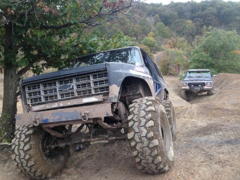 1979 Ford Bronco - Two off-road SUVs navigate a muddy trail, with the lead vehicle a prominent Chevrolet grille and oversized tires.