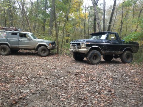 1979 Ford Bronco - Two off-road vehicles, a black Ford Bronco and a grey Jeep Cherokee, are parked on a leaf-covered path in a wooded area.